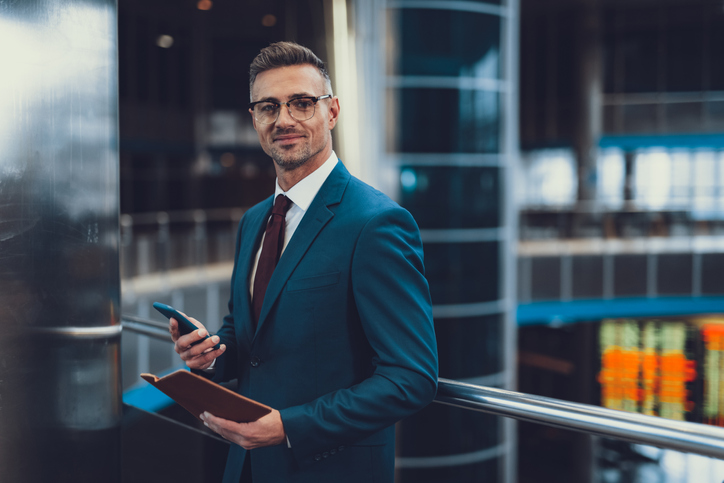 Homem com roupa formal chique em pé dentro do centro de negócios. Ele olhando para a câmera e segurando o telefone com o documento nas mãos. Ele é um investidor anjo em busca de startups.