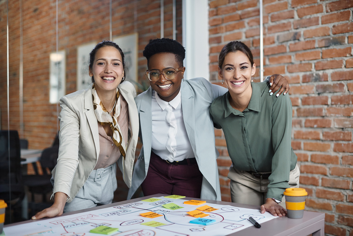 Equipe multirracial de empresárias felizes abraçando enquanto trabalhava em um novo plano de negócio no escritório e olhando para a câmera.