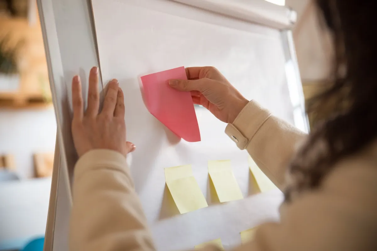 Mãos posicionando uma nota adesiva rosa em um quadro branco com outras notas amarelas, representando organização de tarefas e planejamento visual.