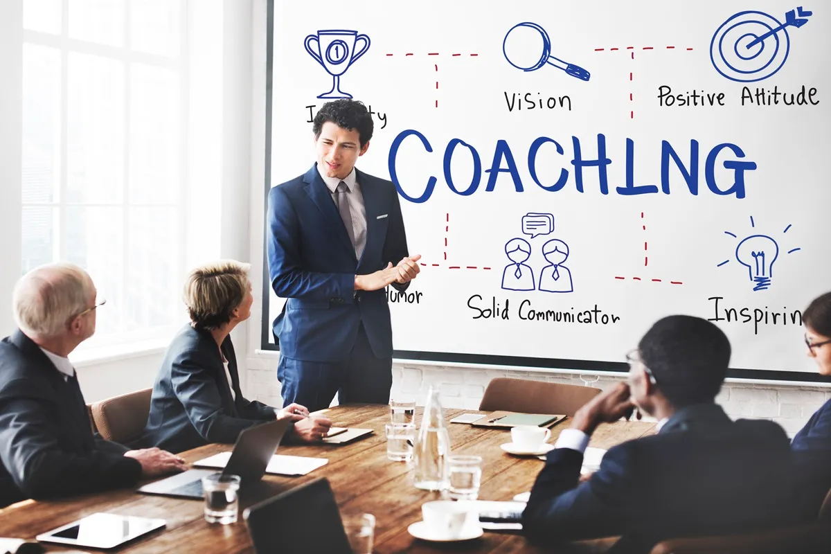 Homem apresentando conceitos de coaching em um quadro branco para uma equipe em reunião.