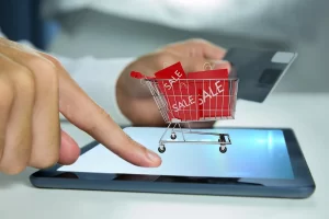 Mão interagindo com tablet exibindo carrinho de compras virtual contendo sacolas vermelhas de promoção e cartão de crédito ao fundo.