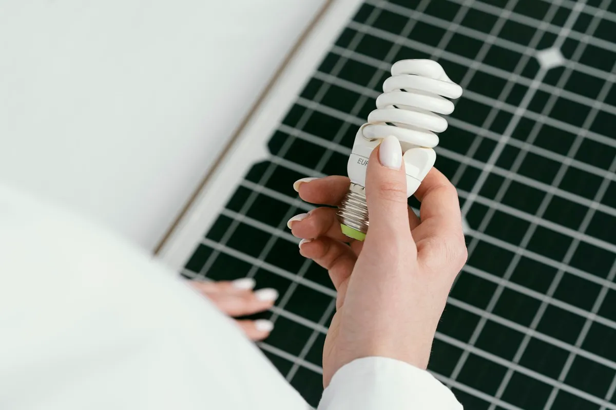 Mão segurando uma lâmpada fluorescente compacta sobre um painel solar, representando eficiência energética e uso de energia renovável.