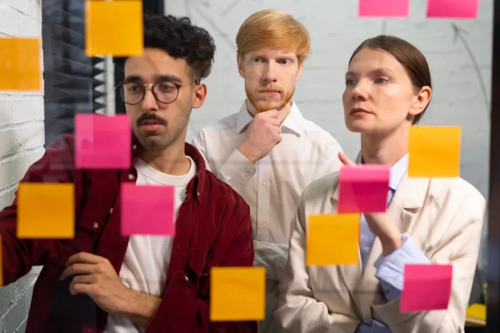 Três profissionais observando e organizando notas adesivas coloridas em um painel de vidro, simbolizando trabalho em equipe e planejamento estratégico