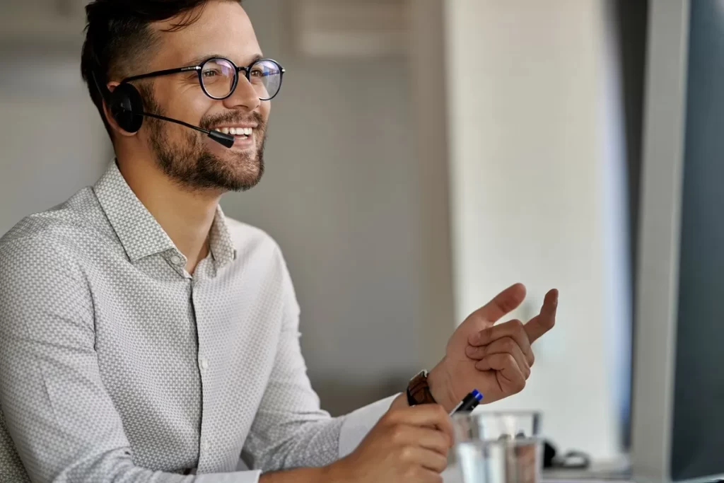 Homem com óculos e headset sorrindo enquanto trabalha no atendimento ao cliente, em frente ao computador.