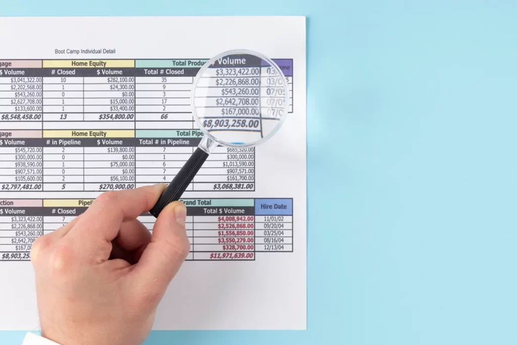 Mão segurando uma lupa sobre uma tabela financeira, ampliando os números para análise detalhada, com fundo azul claro.