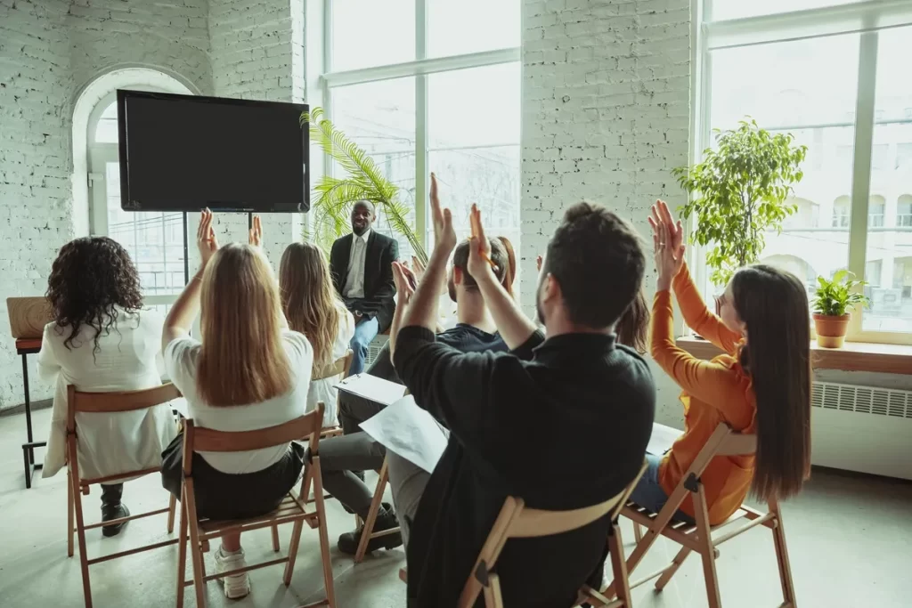 Grupo de pessoas participando de uma sessão de coaching, levantando as mãos enquanto interagem com o palestrante.