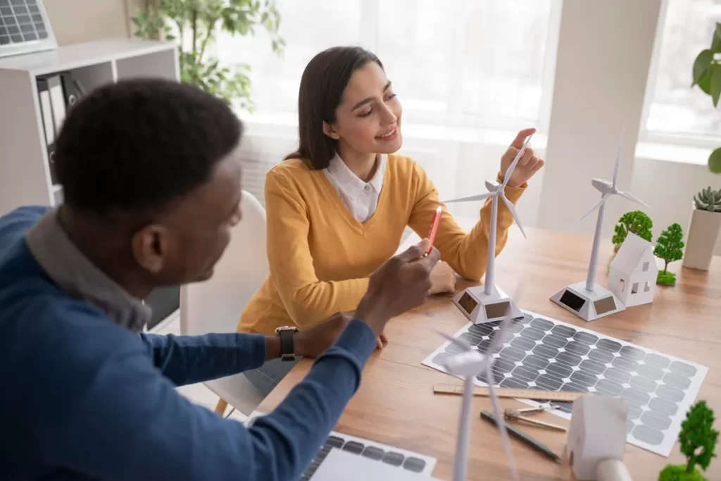 Dois profissionais discutindo projetos de energia sustentável em uma mesa com miniaturas de turbinas eólicas, painéis solares e maquetes de casas