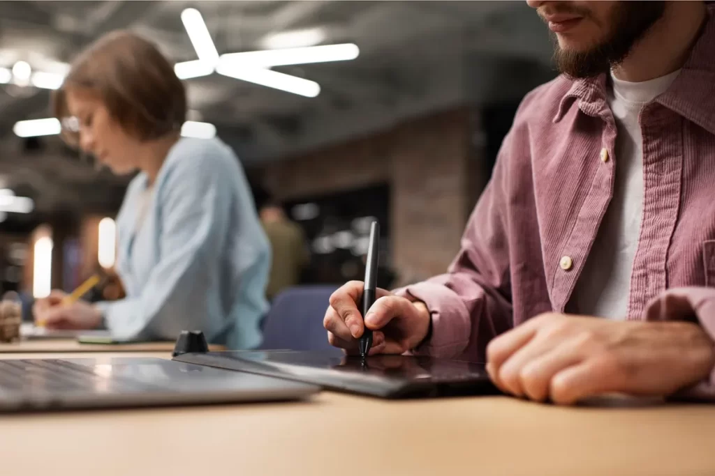 Homem utilizando uma mesa digitalizadora com caneta enquanto trabalha em um ambiente de coworking, com outra pessoa ao fundo escrevendo em um caderno.