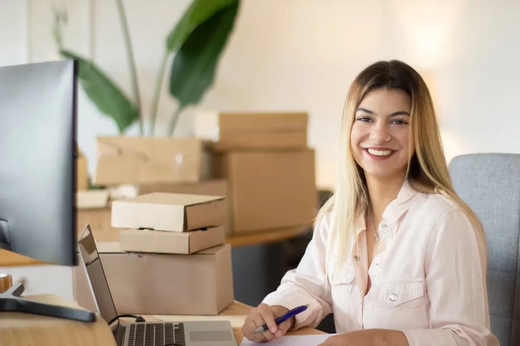 Jovem empreendedora sorridente trabalhando em um ambiente de escritório com caixas empilhadas e computador, representando negócios digitais e modelo de subscrição.