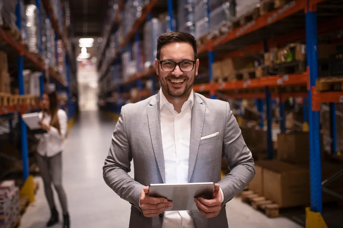 Homem sorridente com óculos e traje social segurando um tablet em um armazém com prateleiras organizadas, representando gestão de logística e controle de inventário